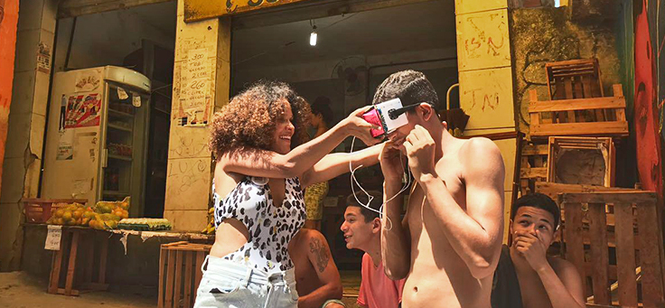 Thamyra está ajudando jovem a colocar em frente aos olhos dispositivo feito com materiais recicláveis para assistir a vídeos 360° por meio de um celular, um exemplo de gambiarra que ajuda a explicar o termo Mecnologia.