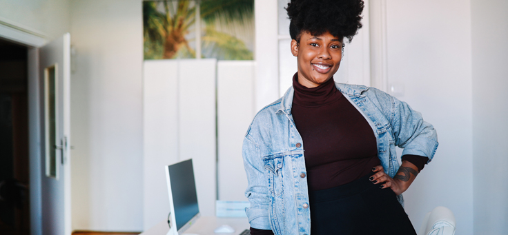 Imagem mostra uma mulher negra empreendedora. Ela usa blusa preta e jaqueta jeans, está com a mão esquerda na cintura. É possível ver um notebook em cima de uma mesa na sala onde a mulher está