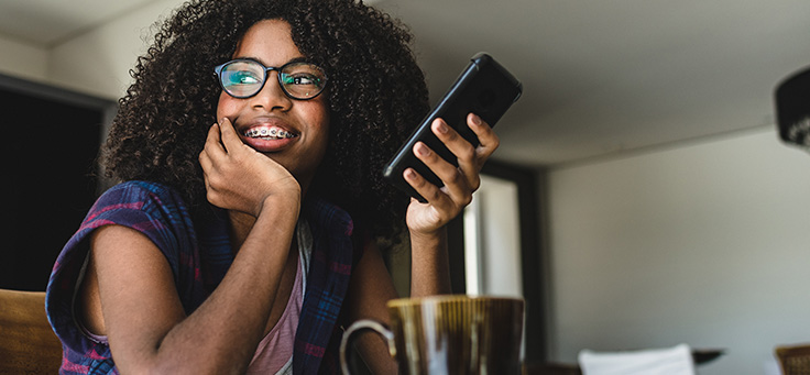 Menina de óculos e cabelos cacheados sorri com o rosto apoiado em uma das mãos e segura um smartphone na outra. Imagem ilustra pauta sobre o Pense Grande Digital, aplicativo que oferece formação gratuita em empreendedorismo social.