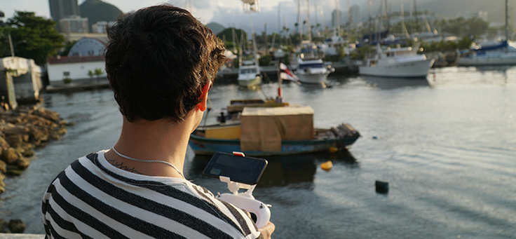 Jovem do projeto Barkus está de costas para a câmera e segura um suporte com um smartphone acoplado. Ele está em uma marina e tem barcos ao fundo da foto