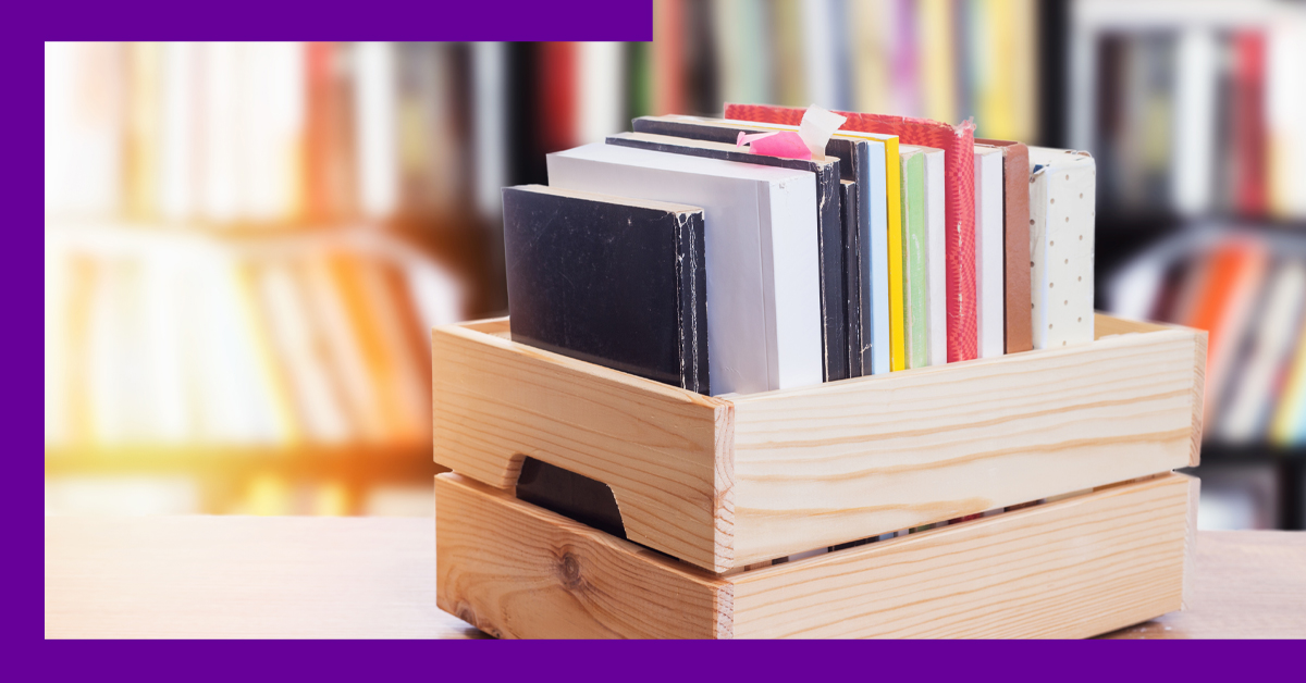 imagem mostra uma caixa de madeira sobre a mesa com diversos livros dentro. Ao fundo, há uma estante de livros.