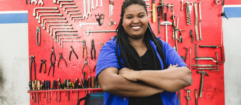 foto mostra uma mulher negra de macacão azul sorrindo para foto. Atrás dela, há uma painel repleto de ferramentas.