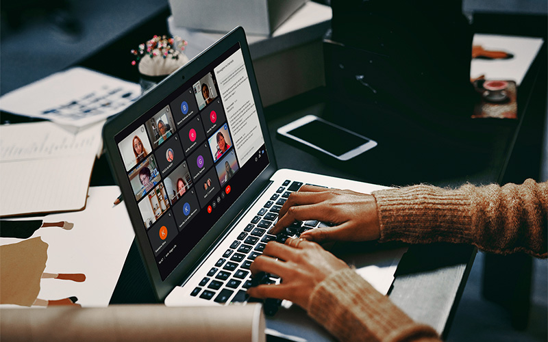 Imagem mostra uma pessoa digitando em um notebook. Na tela do aparelho, é possível ver pessoas participando de um em uma sala virtual. O notebook está sobre uma mesa onde também há um aparelho celular, cadernos e canetas.