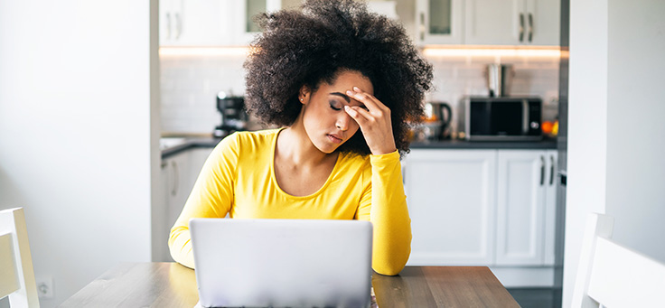 Mulher está sentada em frente a notebook e segurando a testa com uma das mãos em imagem para ilustrar quem sofre da Síndrome do Impostor. Ela é negra, tem os cabelos encaracolados e crespos e está vestindo uma blusa amarela de mangas compridas