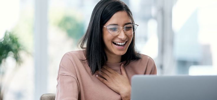 Imagem mostra uma jovem sorrindo. Ela está em frente a um computador. Ela usa óculos e uma blusa na cor rosa.