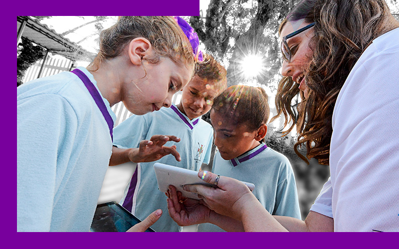 Imagem mostra três crianças olhando e interagindo com um tablet. O aparelho está sendo mostrado por uma professora, que está reunida com os alunos. As crianças estão uniformizadas e todos estão em um ambiente externo.