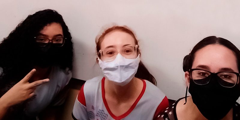 Imagem mostra as três jovens estudantes que desenvolveram aplicativo para ajudar refugiados. As três usam óculos e máscaras de proteção. Todas sorriem para a foto e uma delas está fazendo sinal de “V” com os dedos.