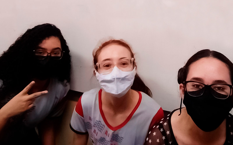 Imagem mostra as três jovens estudantes que desenvolveram aplicativo para ajudar refugiados. As três usam óculos e máscaras de proteção. Todas sorriem para a foto e uma delas está fazendo sinal de “V” com os dedos.