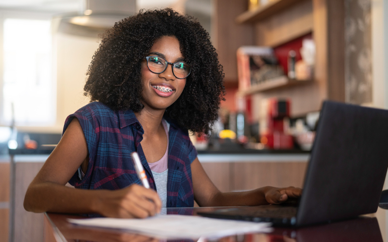 Imagem mostra uma jovem negra em um ambiente que parece ser a sua casa. Ela está sentada, em uma mesa à sua frente há um notebook preto e com a mão direita ela segura uma caneta. Na mesa, também há papéis. A jovem usa óculos, uma blusa branca e um colete xadrez, e sorri para a foto.