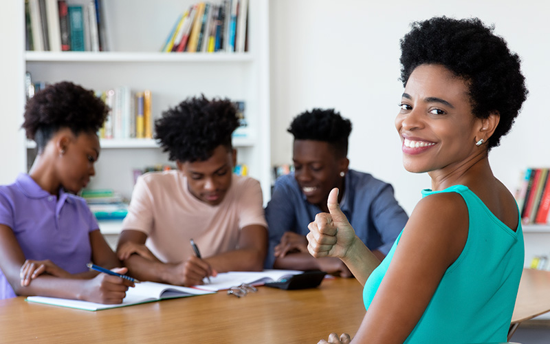 Imagem mostra quatro jovens negros reunidos em uma mesa. Três deles estão de um lado, observando algo que está sendo escrito por um deles em um caderno. A outra jovem, que pode ser a professora dos demais, está à frente, fazendo sinal de positivo e sorrindo para a foto.