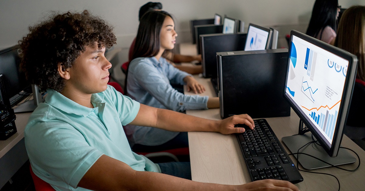 Imagem mostra três jovens em uma sala de aula, utilizando computadores. Em primeiro plano, é possível ver um jovem rapaz, de cabelo cacheado, e ao seu lado uma moça, de cabelo comprido.