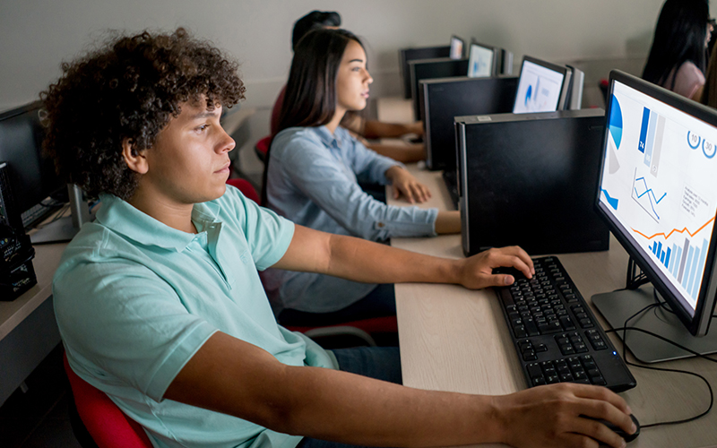 Imagem mostra três jovens em uma sala de aula, utilizando computadores. Em primeiro plano, é possível ver um jovem rapaz, de cabelo cacheado, e ao seu lado uma moça, de cabelo comprido.