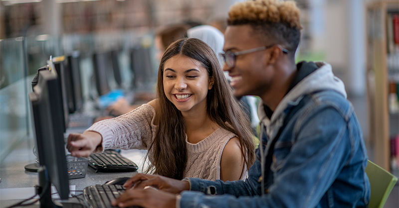 Imagem mostra uma jovem branca e um jovem negro em uma sala de aula com computadores. Os dois estão juntos; ela sorri e aponta para algo na tela do computador, enquanto ele também sorri e observa a tela do aparelho.