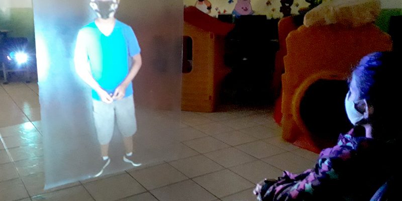 Imagem mostra a holografia de um aluno sendo projetada em sala de aula. A holografia foi criada pelo professor Alberto Santos. Uma aluna está sentada assistindo à exibição da holografia.