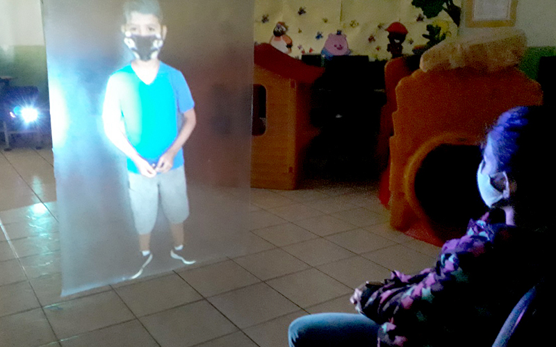 Imagem mostra a holografia de um aluno sendo projetada em sala de aula. A holografia foi criada pelo professor Alberto Santos. Uma aluna está sentada assistindo à exibição da holografia.