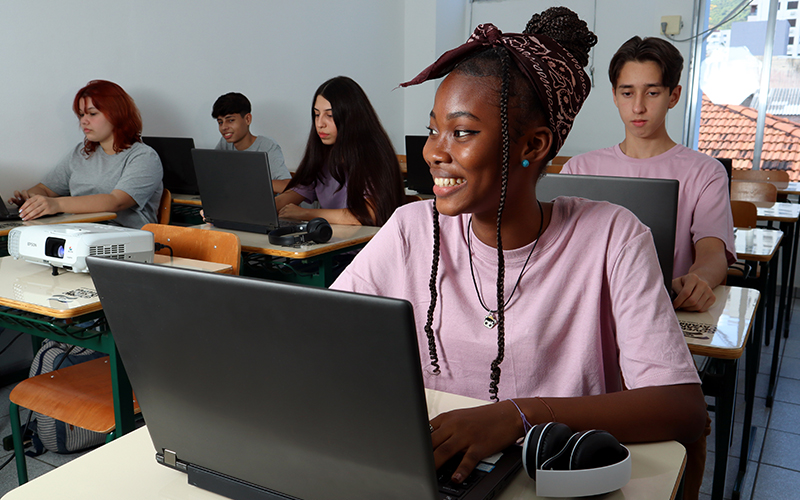 Imagem mostra uma sala de aula onde há seis estudantes. Todos estão sentados em carteiras, utilizando notebooks. Em primeiro plano, há uma jovem negra, que está sorrindo.