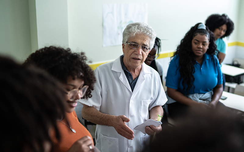 Imagem mostra um professor no centro da imagem. Ele está rodeado por cinco estudantes. Além disso, segura um papel com uma das mãos. Ele parece estar falando algo para os alunos. Todos estão em uma sala de aula.