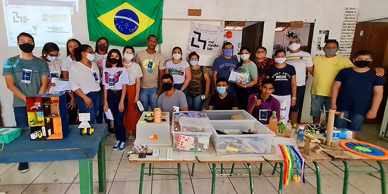 Imagem mostra um grupo de 20 pessoas reunidos em um ambiente onde há mesas e objetos pedagógicos sobre elas. Ao fundo, uma bandeira do Brasil e uma imagem de projetor refletida na parede. O grupo é formado por homens, mulheres e jovens.