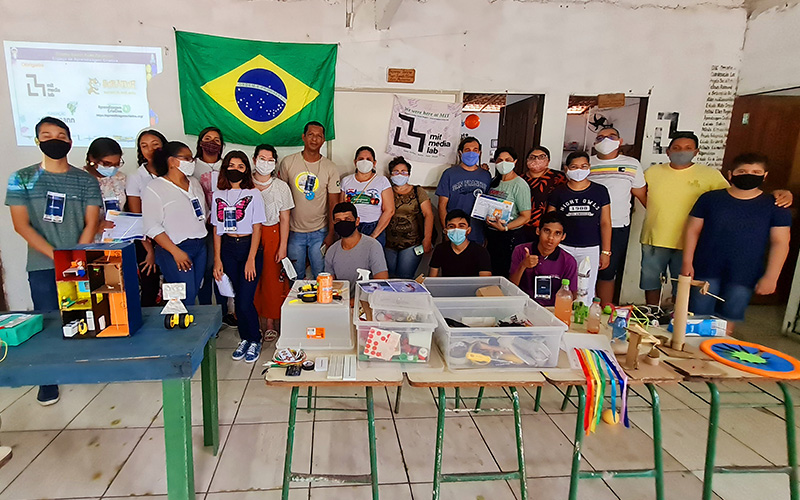 Imagem mostra um grupo de 20 pessoas reunidos em um ambiente onde há mesas e objetos pedagógicos sobre elas. Ao fundo, uma bandeira do Brasil e uma imagem de projetor refletida na parede. O grupo é formado por homens, mulheres e jovens.