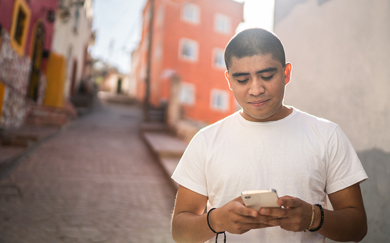 Jovem está mexendo em um aparelho de celular enquanto caminha por uma rua dentro de uma comunidade na América Latina. Ele é moreno, tem os cabelos curtos e usa uma camiseta branca.