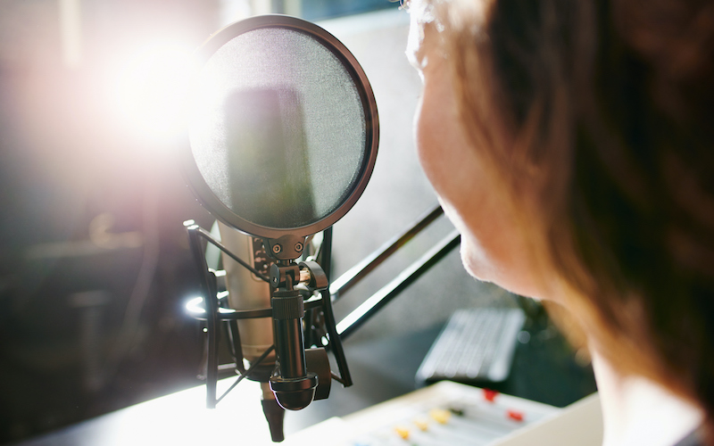 Mulher de costas, falando ao microfone, gravando um áudio, em ambiente escolar.