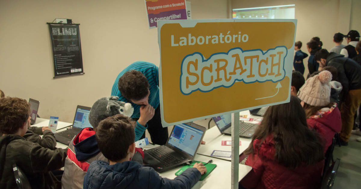 Imagem mostra laboratório de informática onde alunos e professores interagem durante o uso de computadores e da plataforma Scratch. Em primeiro plano há uma placa laranja com os dizeres 