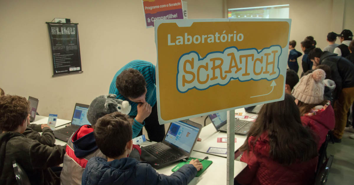 Imagem mostra laboratório de informática onde alunos e professores interagem durante o uso de computadores e da plataforma Scratch. Em primeiro plano há uma placa laranja com os dizeres