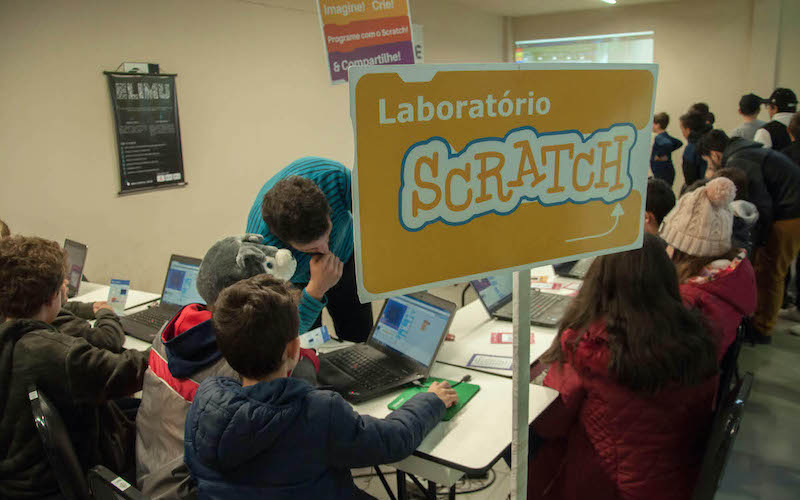 Imagem mostra laboratório de informática onde alunos e professores interagem durante o uso de computadores e da plataforma Scratch. Em primeiro plano há uma placa laranja com os dizeres "Laboratório Scratch".