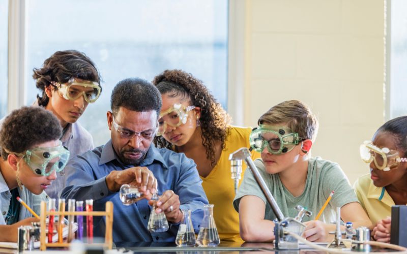 Imagem mostra professor e alunos reunidos debatendo e, ao mesmo tempo, realizando um experimento em laboratório.