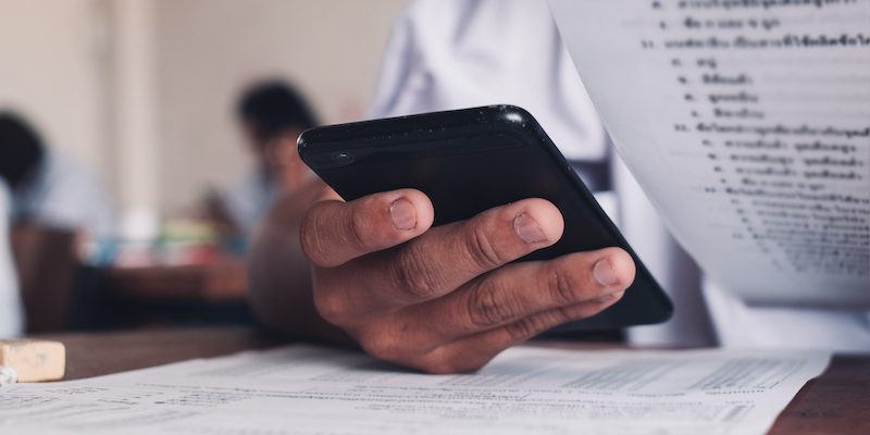 Mão segurando um celular em uma sala de aula, junto com outros materiais educativos.