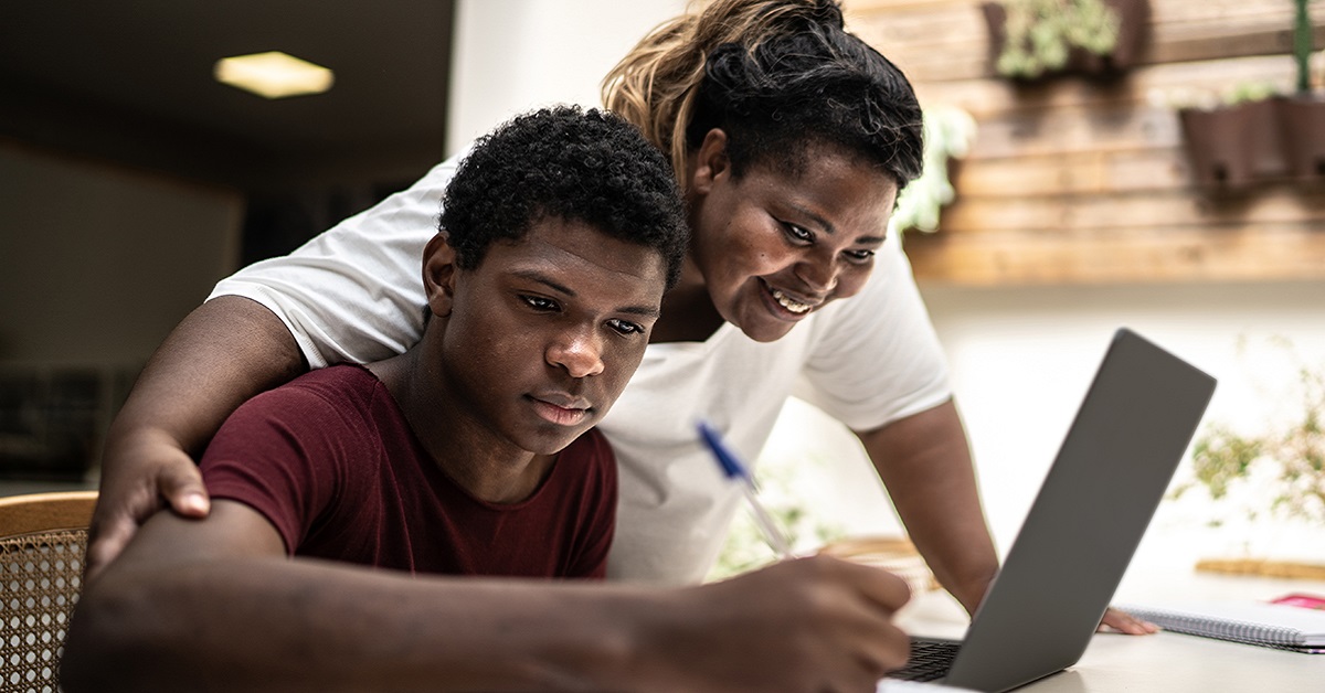 Imagem mostra um jovem estudante negro em um ambiente que parece ser sua casa. Ele está estudando, utilizando um notebook. Sua mãe está ao seu lado oferecendo apoio a ele.
