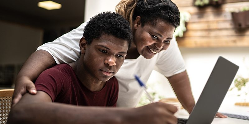 Imagem mostra um jovem estudante negro em um ambiente que parece ser sua casa. Ele está estudando, utilizando um notebook. Sua mãe está ao seu lado oferecendo apoio a ele.