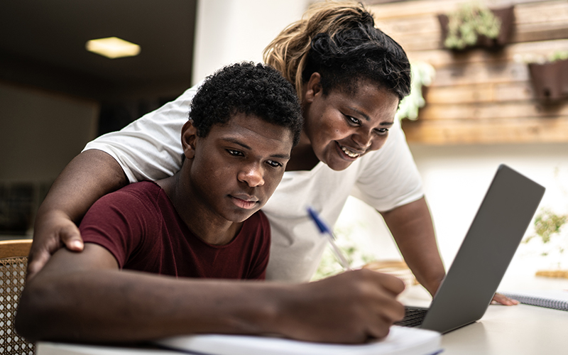 Imagem mostra um jovem estudante negro em um ambiente que parece ser sua casa. Ele está estudando, utilizando um notebook. Sua mãe está ao seu lado oferecendo apoio a ele.