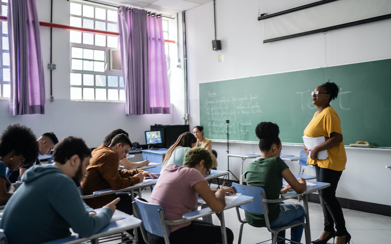 Sala de aula com professor de pé, alunos sentados na carteira fazendo anotações e lousa verde.