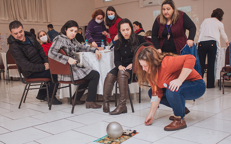 Imagem mostra um grupo de professores (homens e mulheres) participando de uma dinâmica durante o evento. Uma professora está interagindo com uma bola no chão, enquanto os demais professores observam sua ação, alguns professores estão sentados e outros estão em pé.