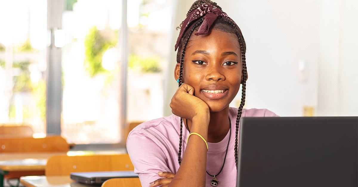 Imagem mostra uma menina negra em uma sala de aula. ela sorri para a foto, está sentada em frente a um computador e usa camiseta rosa