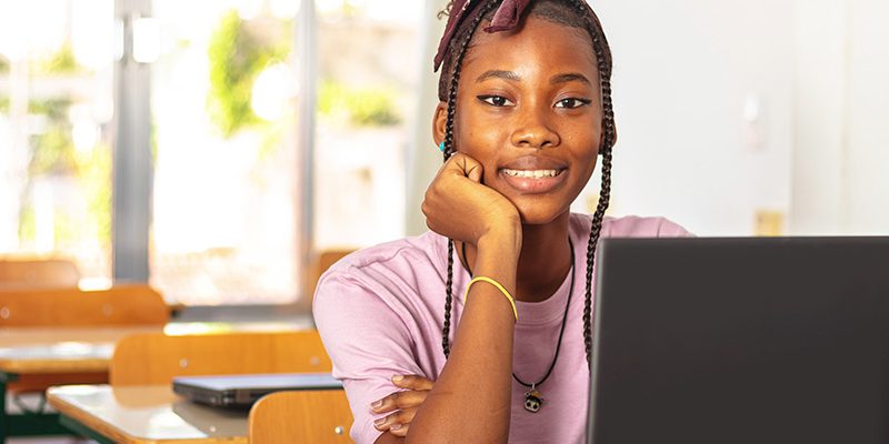 Imagem mostra uma menina negra em uma sala de aula. ela sorri para a foto, está sentada em frente a um computador e usa camiseta rosa