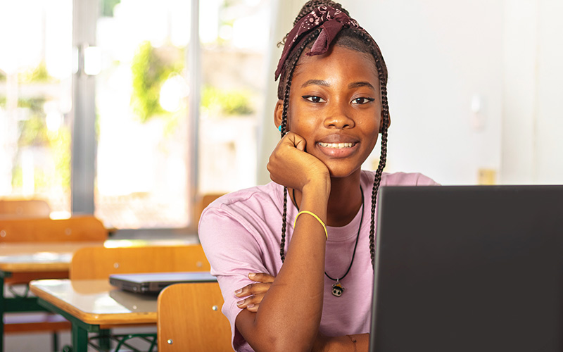 Imagem mostra uma menina negra em uma sala de aula. ela sorri para a foto, está sentada em frente a um computador e usa camiseta rosa