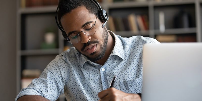 Professor utiliza óculos e fone em frente ao computador, enquanto faz anotações com caneta em um caderno.