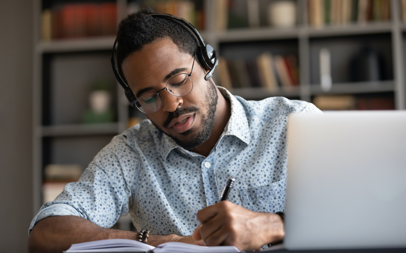 Professor utiliza óculos e fone em frente ao computador, enquanto faz anotações com caneta em um caderno.