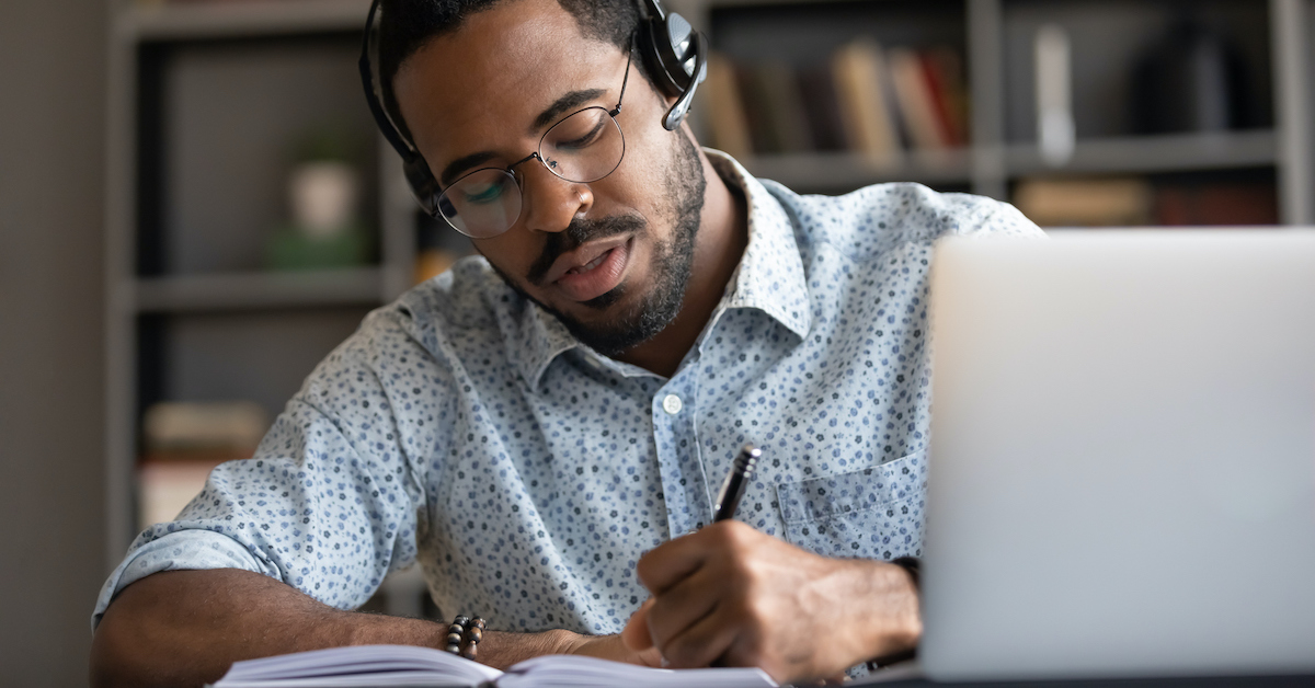 Professor utiliza óculos e fone em frente ao computador, enquanto faz anotações com caneta em um caderno.
