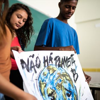 Imagem mostra uma adolescente branca e um adolescente negro segurando um cartaz com a pintura de um globo terrestre e com a frase Não há planeta. Eles estão em uma sala de aula.