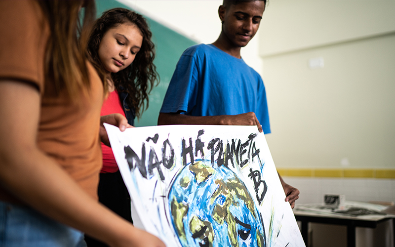 Imagem mostra uma adolescente branca e um adolescente negro segurando um cartaz com a pintura de um globo terrestre e com a frase Não há planeta. Eles estão em uma sala de aula.