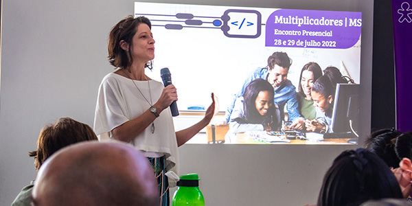 Imagem mostra Lia Roitburd apresentando a formação aos educadores. Ao fundo, uma imagem é projetada em uma tela. Lia tem cabelos curtos, usa camisa branca e segura um microfone