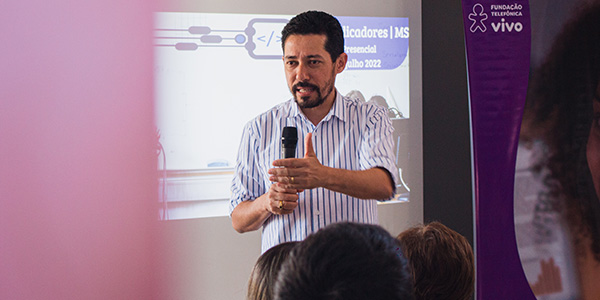 Imagem mostra professor Davi de Oliveira falando aos educadores presentes. Ele é moreno, usa barba, veste camisa branca listrada e segura um microfone