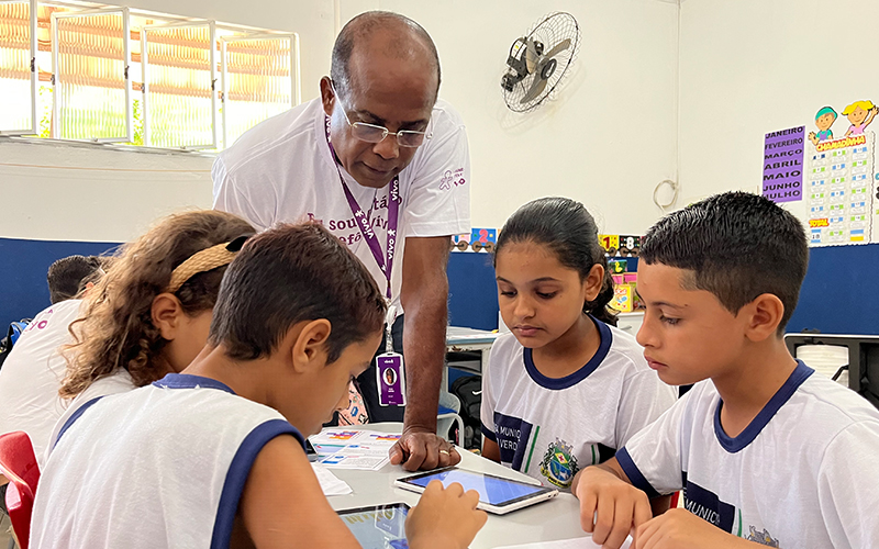Imagem mostra um voluntário negro orientando quatro alunos em uma sala de aula. O voluntário usa óculos. Os alunos estão sentados e observam tablets que estão em cima da mesa.