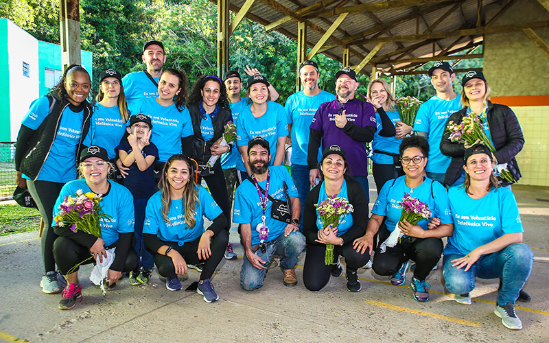 Imagem mostra grupo de 19 voluntários, entre homens e mulheres, reunidos em uma instituição. Eles usam camiseta azul com a frase Eu sou voluntário Telefônica