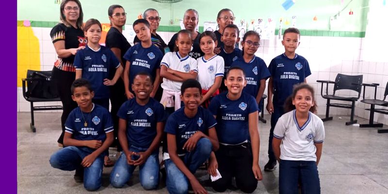 Na imagem, vemos um grupo de 12 crianças que são alunos monitores do Aula Digital no Sergipe, em uma sala de aula reunidos com cinco professoras
