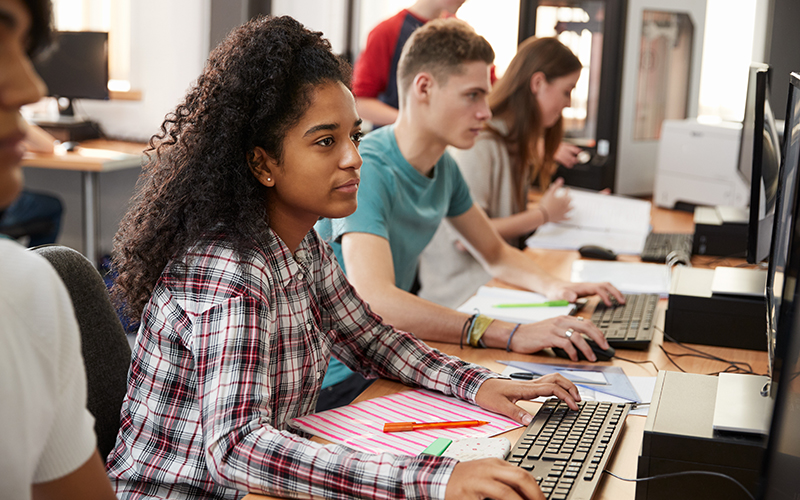 Imagem mostra três jovens em uma sala de aula utilizando computadores. Em primeiro plano há uma jovem negra e ao se lado um jovem e uma jovem branca