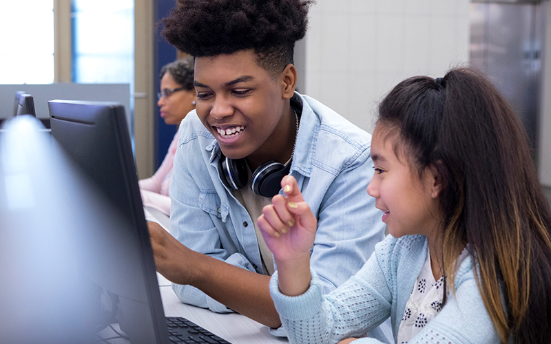 Imagem mostra um adolescente negro e uma adolescente com traços orientais juntos olhando para a tela de um computador. Ao fundo, há uma mulher negra usando um computador